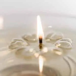 A close-up of a small Aromaglow floating candle lit with a single wick, surrounded by soft ripples and reflections in a light-colored surface.