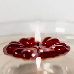 A red Aromaglow floating candle in the shape of a flower, lit with a small flame in the center, set against a soft, out-of-focus light background.