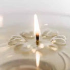 A close-up of a small Aromaglow floating candle lit with a single wick, surrounded by soft ripples and reflections in a light-colored surface.