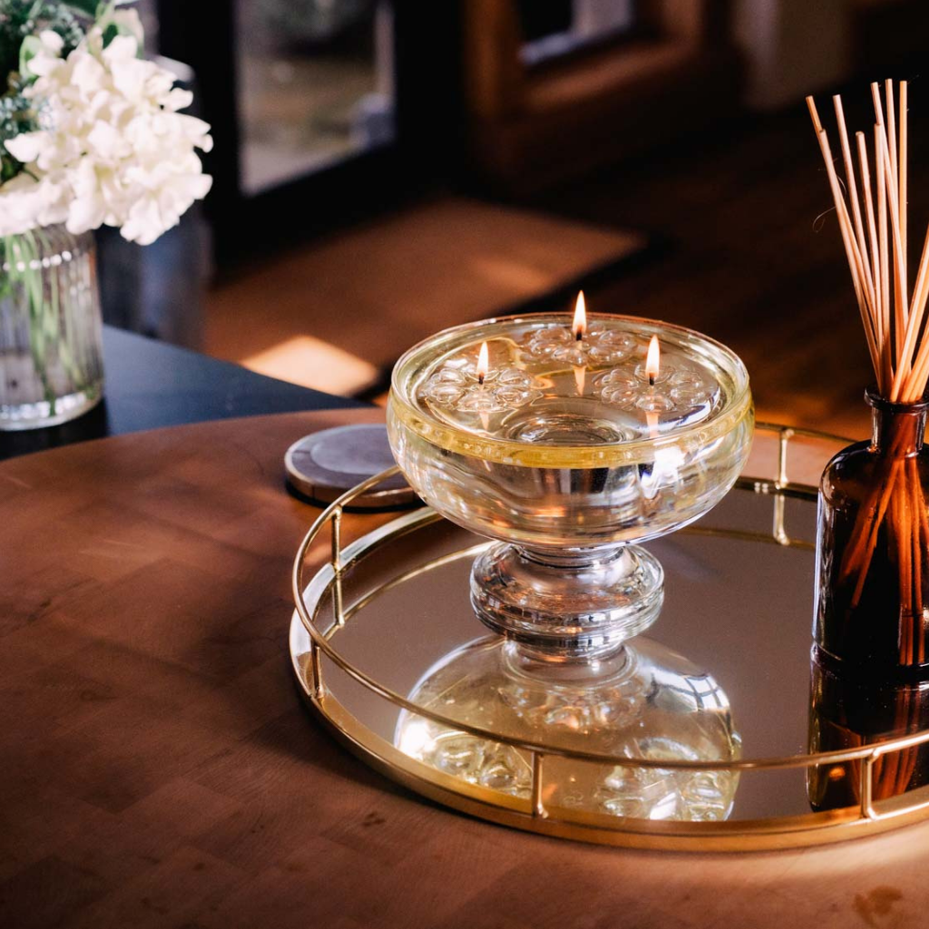 A clear glass candle holder filled with water has three lit Aromaglow floating candles on a round mirrored tray next to a brown reed diffuser bottle with wooden sticks, set on a warm wood table in a softly lit interior.