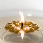 A close-up of an Aromaglow floating gold candle with a warm yellow flame, showing the translucent golden oil inside a clear circular holder on a light background.