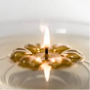 A close-up of an Aromaglow floating gold candle with a warm yellow flame, showing the translucent golden oil inside a clear circular holder on a light background.