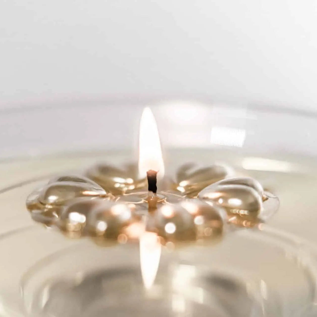 A close-up of an Aromaglow floating candle in a metallic gold flower-shaped wax float and a lit flame, set in clear glass with a soft, neutral background.