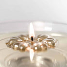 A close-up of an Aromaglow floating candle in a metallic gold flower-shaped wax float and a lit flame, set in clear glass with a soft, neutral background.
